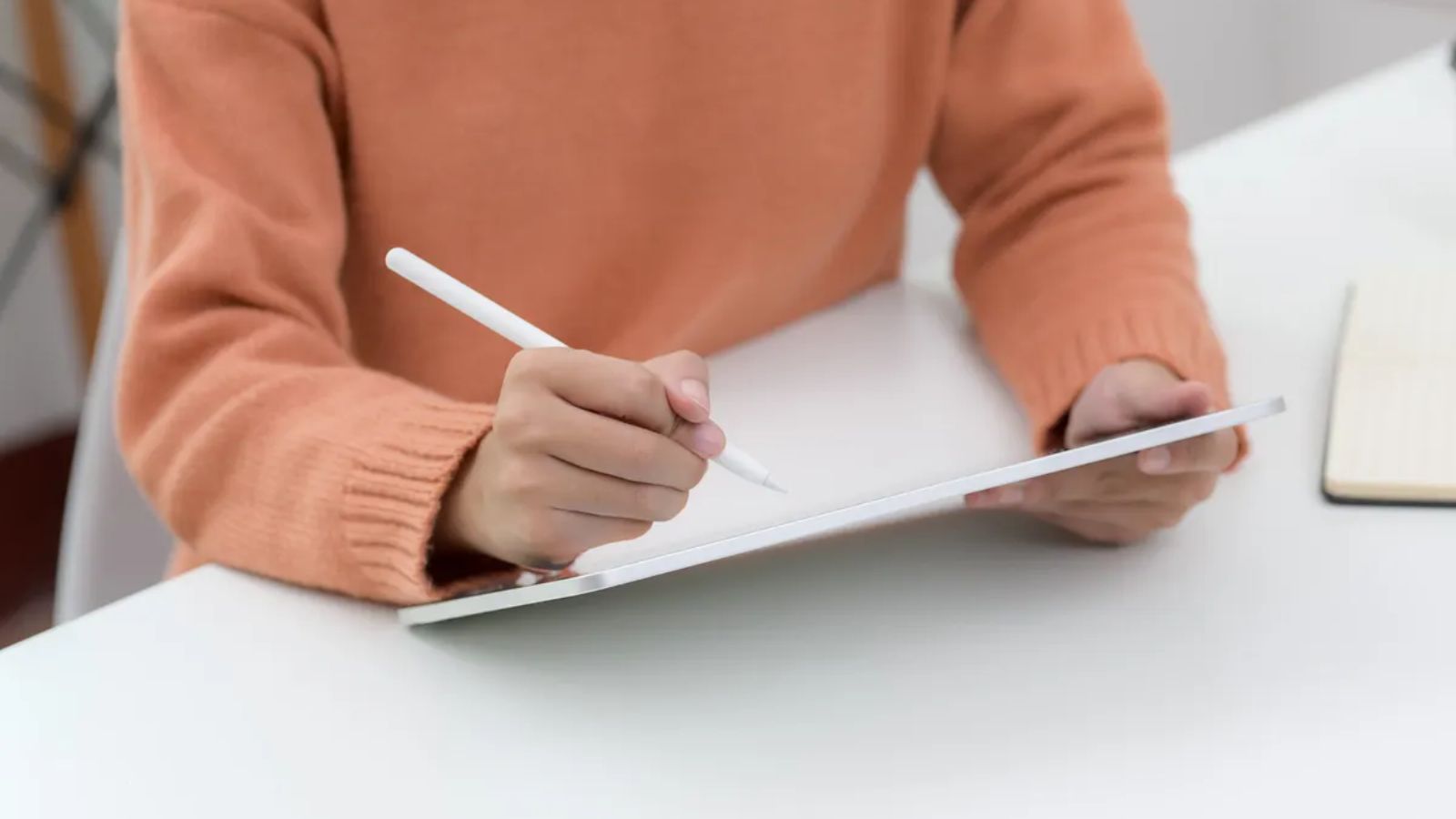 Person writing notes on a tablet with a stylus, notebook on the desk beside them